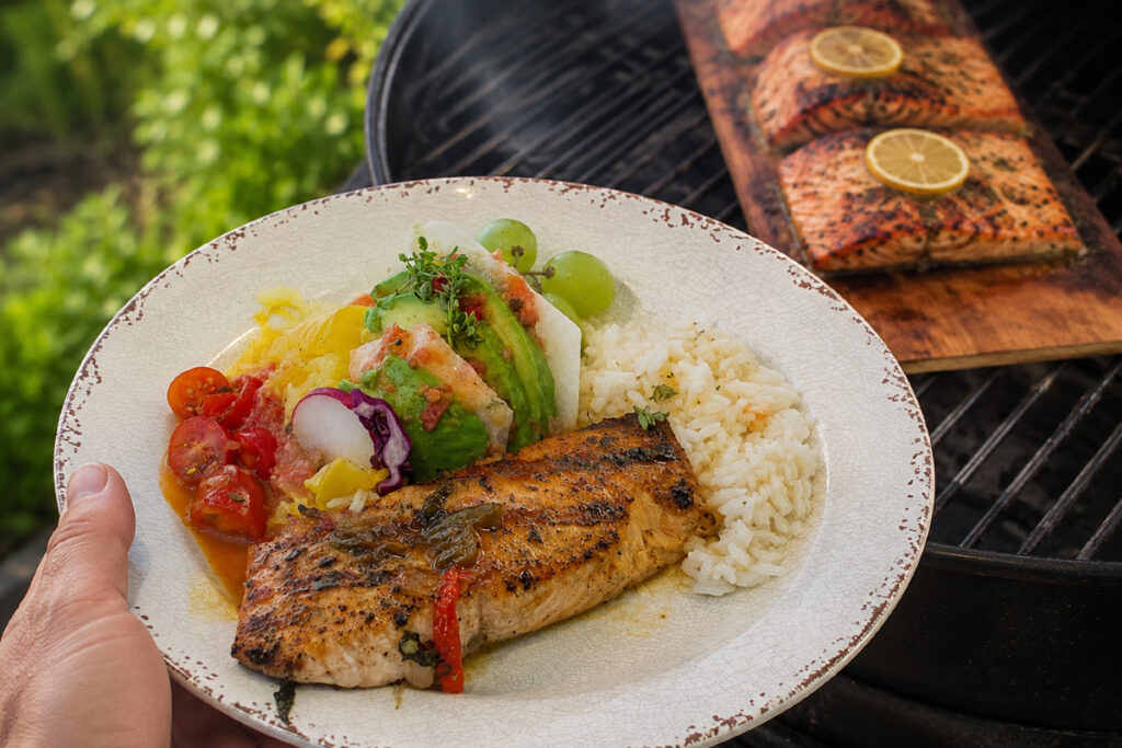 hand holding plate of food with fish, rice, veggeis and grapes with BBQ grill in the background.